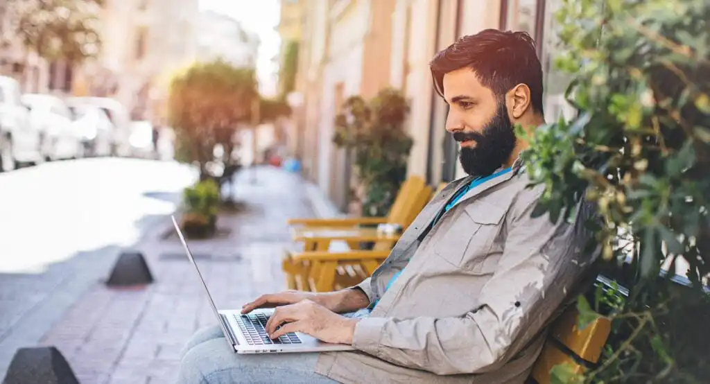 A man with a beard sits on an outdoor bench near greenery, working on a laptop. The sunny street scene around him is calm and inviting.