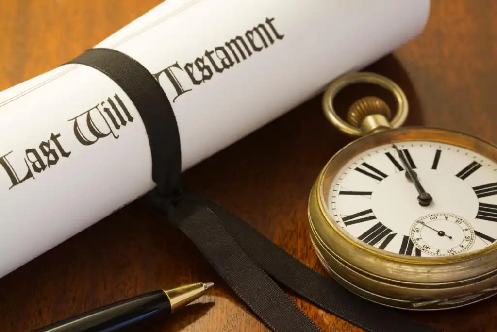 A rolled-up document labeled Last Will Testament tied with a black ribbon, a vintage pocket watch showing 3:45, and a black and gold pen on a wooden surface.