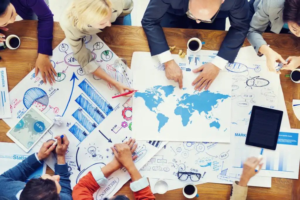 A group of people sit around a table covered with charts, graphs, and a world map, collaborating and discussing data, with coffee cups, pens, and a tablet visible among the papers.