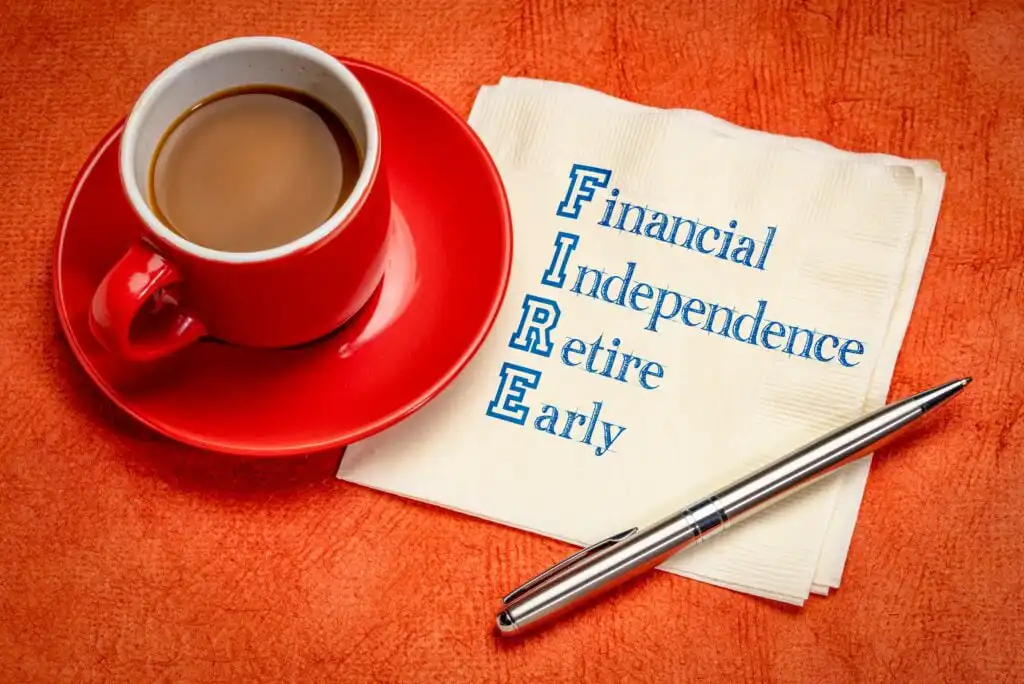 A red coffee cup on a saucer sits next to a napkin with “FIRE: Financial Independence Retire Early” printed on it, alongside a silver pen, all on an orange textured surface.