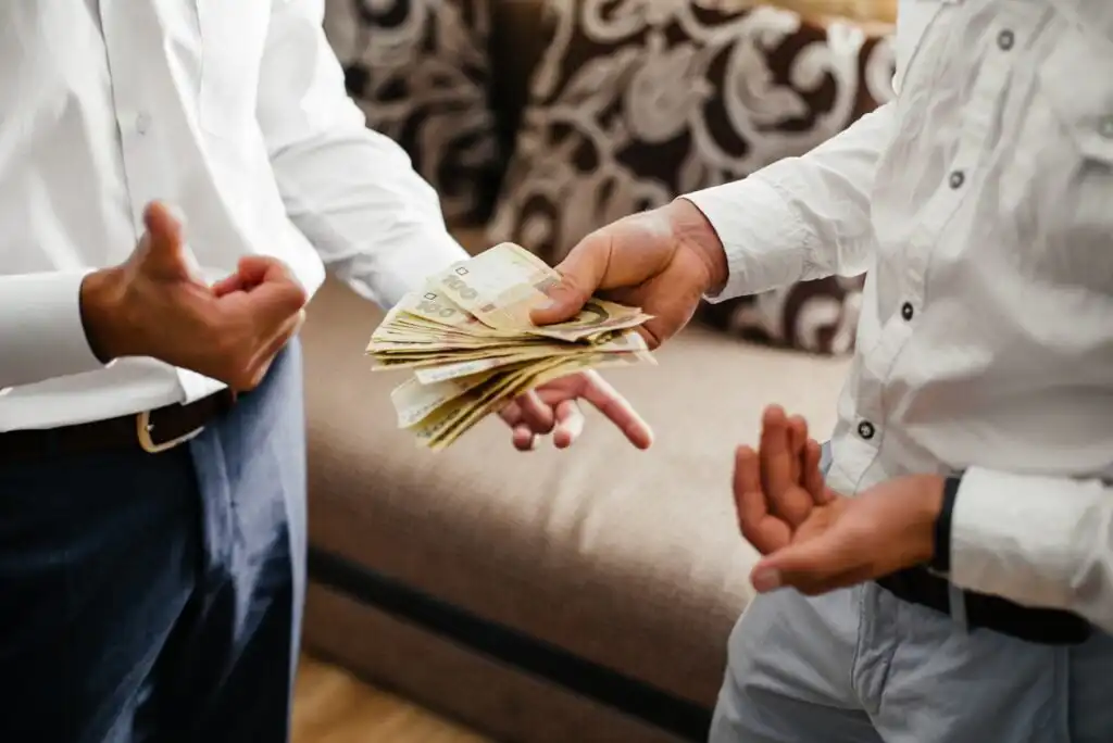Two people standing indoors, one handing a stack of paper currency to the other. Both are dressed in white shirts; only their torsos and hands are visible. A patterned sofa is in the background.