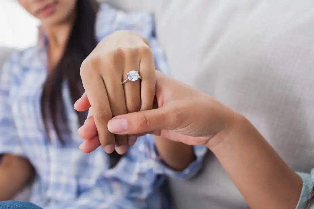 A close-up of one person holding another’s hand, displaying a large diamond engagement ring. Both are sitting on a couch, with one person wearing a blue plaid shirt in the background.