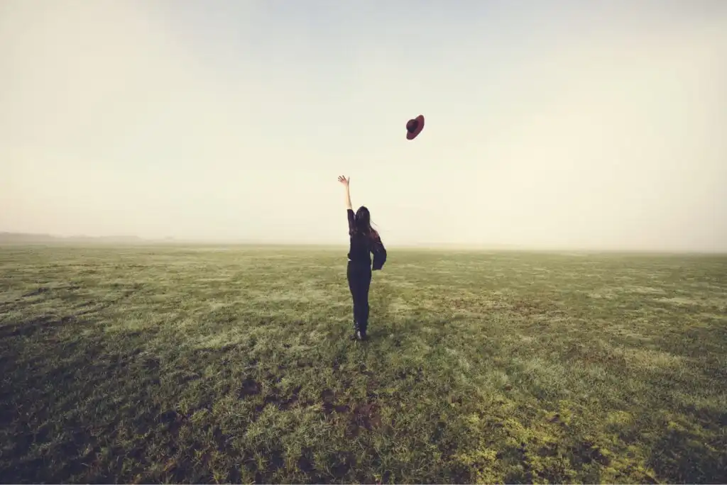 A person stands in a wide, open grassy field under a cloudy sky, facing away while tossing a hat into the air with one hand raised. The scene feels peaceful and carefree.