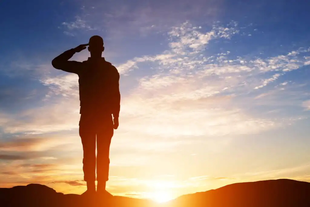 Silhouette of a person in military uniform saluting against a colorful sky at sunrise or sunset, with the sun low on the horizon and scattered clouds above.