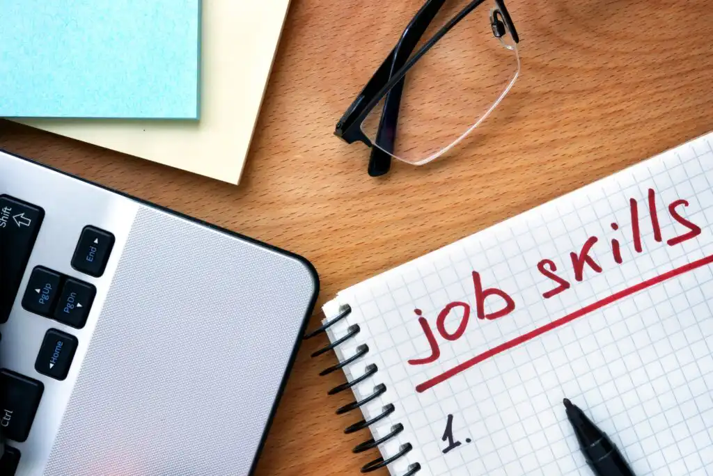 A notepad with job skills written in red marker lies on a desk beside a laptop keyboard, a pen, a pair of glasses, and some sticky notes.
