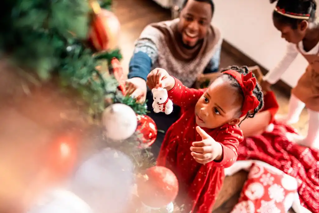A young girl in a red dress decorates a Christmas tree with a white ornament, while an adult and another child watch and smile in the background. The scene is festive and joyful.