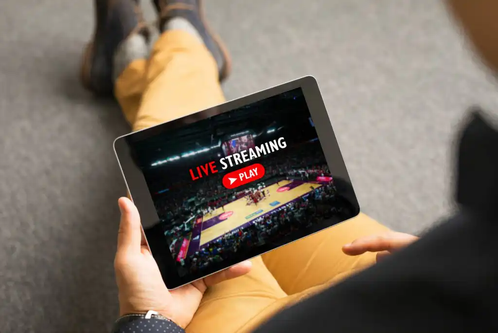 A person sitting on the floor and holding a tablet, watching a live stream of a basketball game, with a “LIVE STREAMING” play button displayed on the screen.