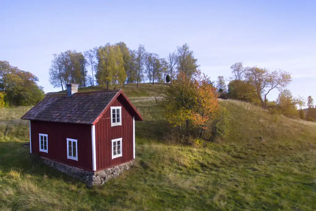 A small red wooden house with white trim sits on a grassy hillside, surrounded by trees with early autumn foliage under a clear blue sky.