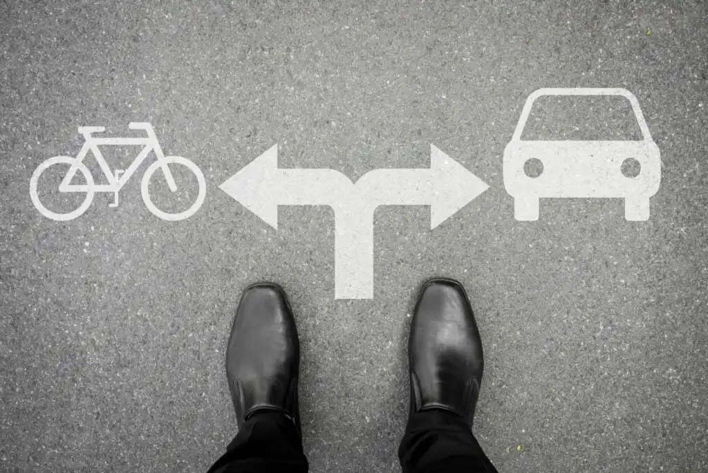A person in black shoes stands on pavement with painted white arrows pointing left to a bicycle symbol and right to a car symbol, suggesting a choice between cycling and driving.