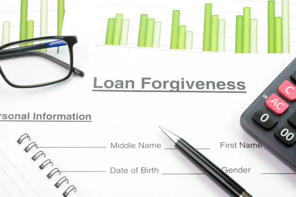A document titled Loan Forgiveness lies on a desk with a pen, eyeglasses, a calculator, and a chart with green bars. There are blank fields for personal information such as name and date of birth.