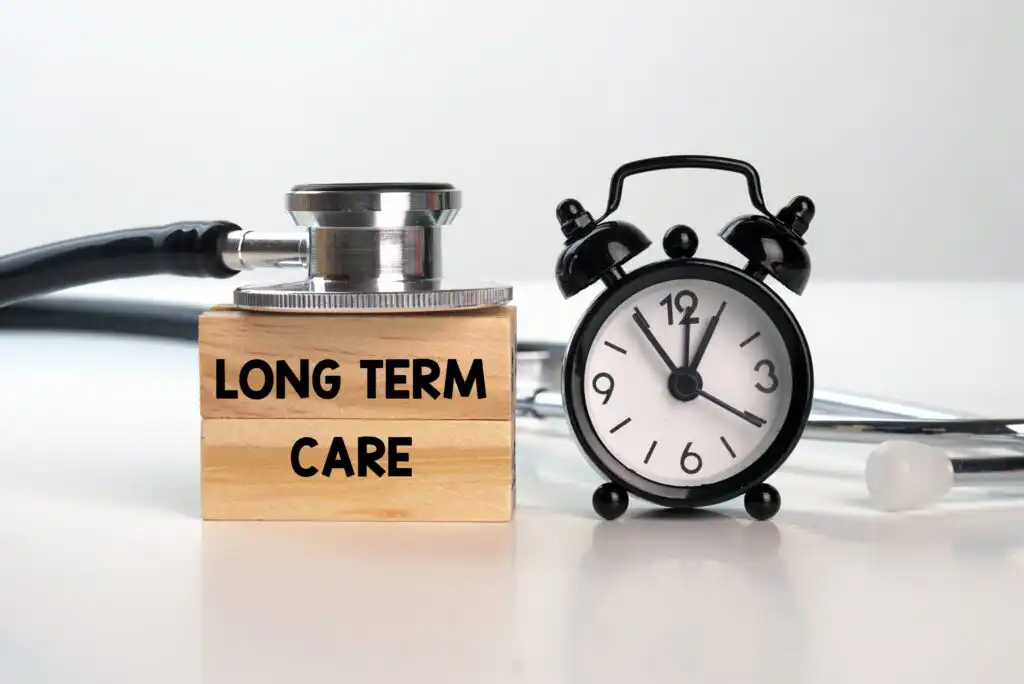 A stethoscope, a wooden block labeled LONG TERM CARE, and a small black alarm clock are arranged together on a white surface, symbolizing healthcare and time.