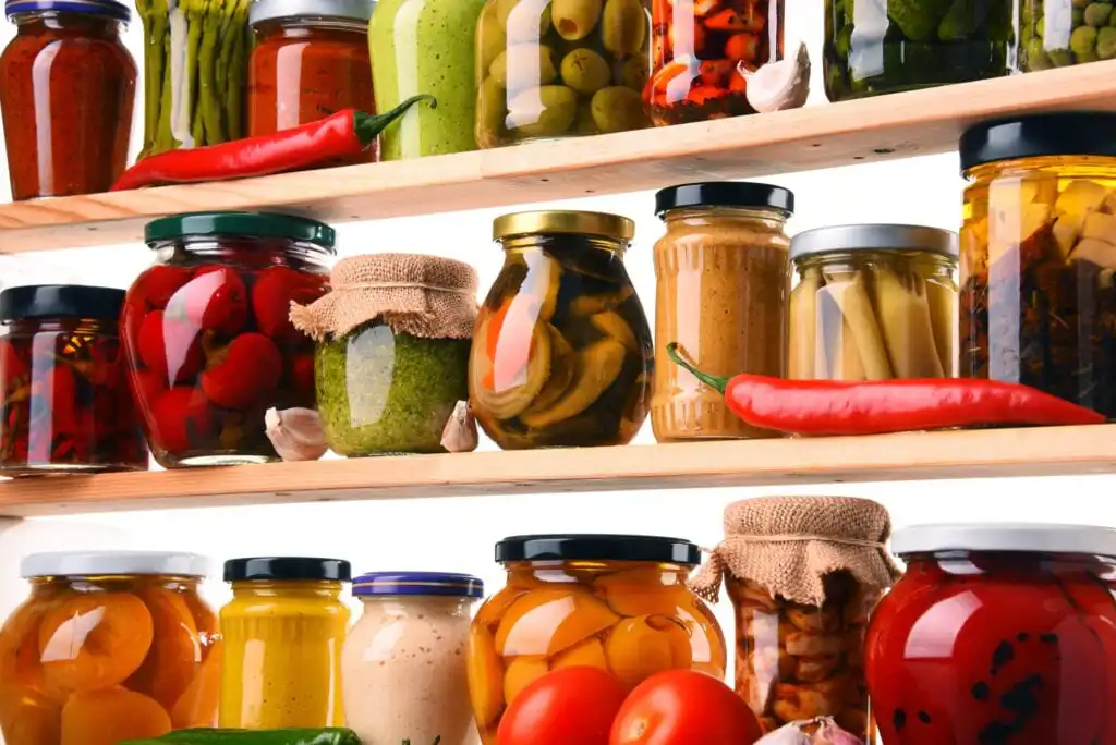 Jars of preserved vegetables, pickles, sauces, and jams in glass containers are arranged on wooden shelves. Bright red chili peppers and tomatoes are placed in front. The scene suggests a pantry or kitchen storage.