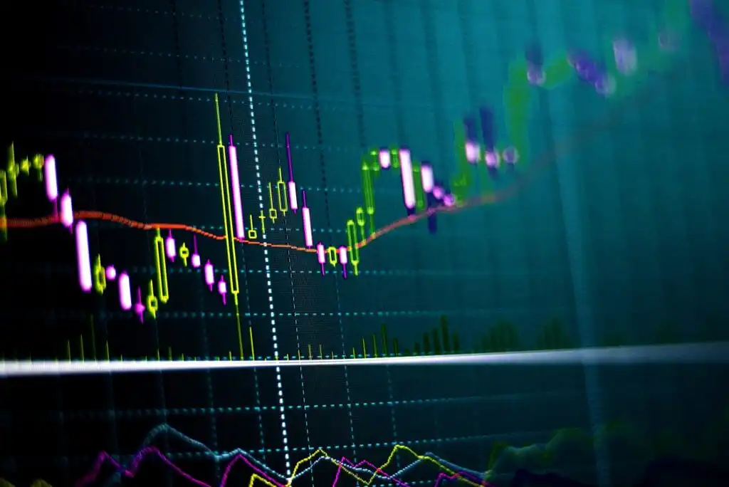 A digital display showing colorful candlestick and line charts, representing financial or stock market data trends, with various indicators against a dark, grid-lined background.