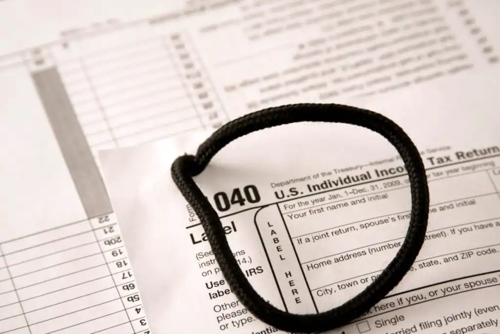 A close-up of U.S. 1040 tax forms with a black hair tie resting on top, placed on a desk. The forms contain various fields for personal and financial information.