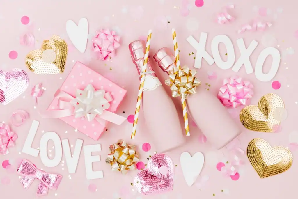 Flat lay of two pink champagne bottles with gold bows, pink gift box, white LOVE and XOXO letters, heart-shaped decorations, and confetti on a pastel pink background.