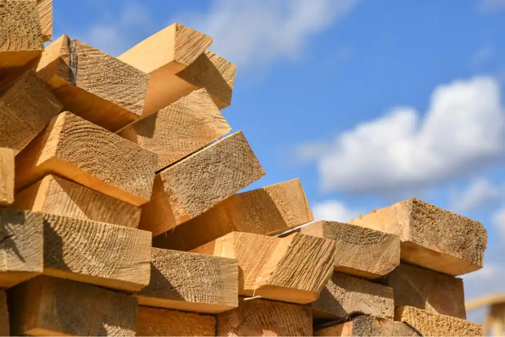 A stack of rough-cut wooden planks is piled outdoors under a blue sky with scattered white clouds. The ends of the lumber face outward, displaying their textured surfaces.