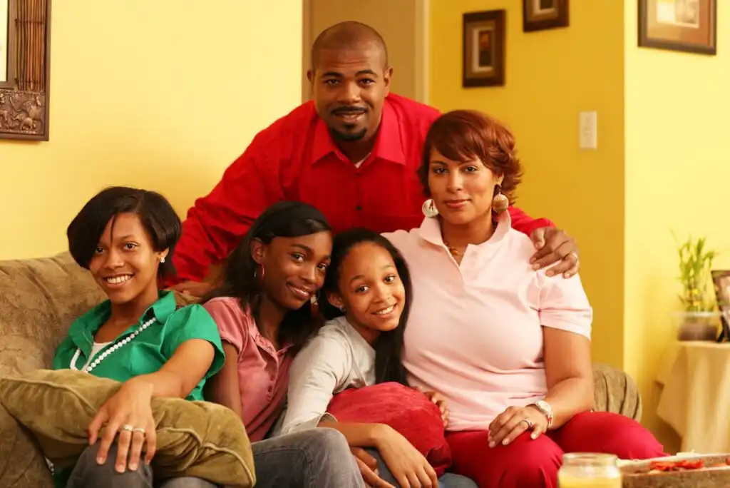 A family of five sits together on a couch, smiling. An adult man stands behind an adult woman and three girls, all sitting close, in a warmly lit living room with yellow walls and framed pictures.