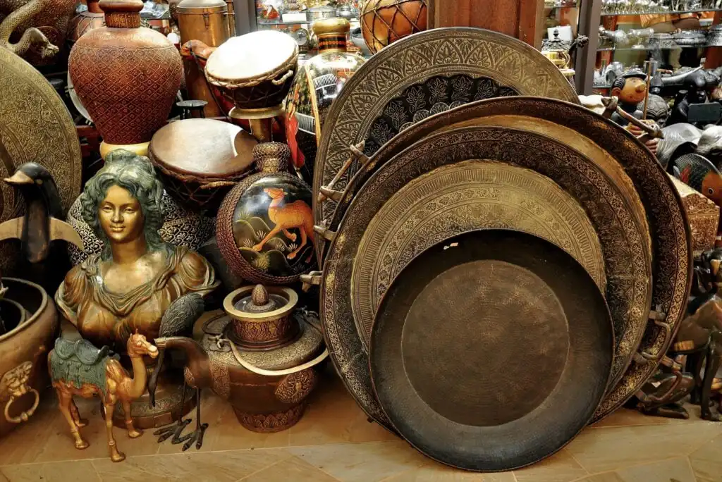 A collection of ornate brass plates, vases, drums, and pottery, with an antique bust of a woman and a camel figurine, displayed together in a shop filled with decorative items.