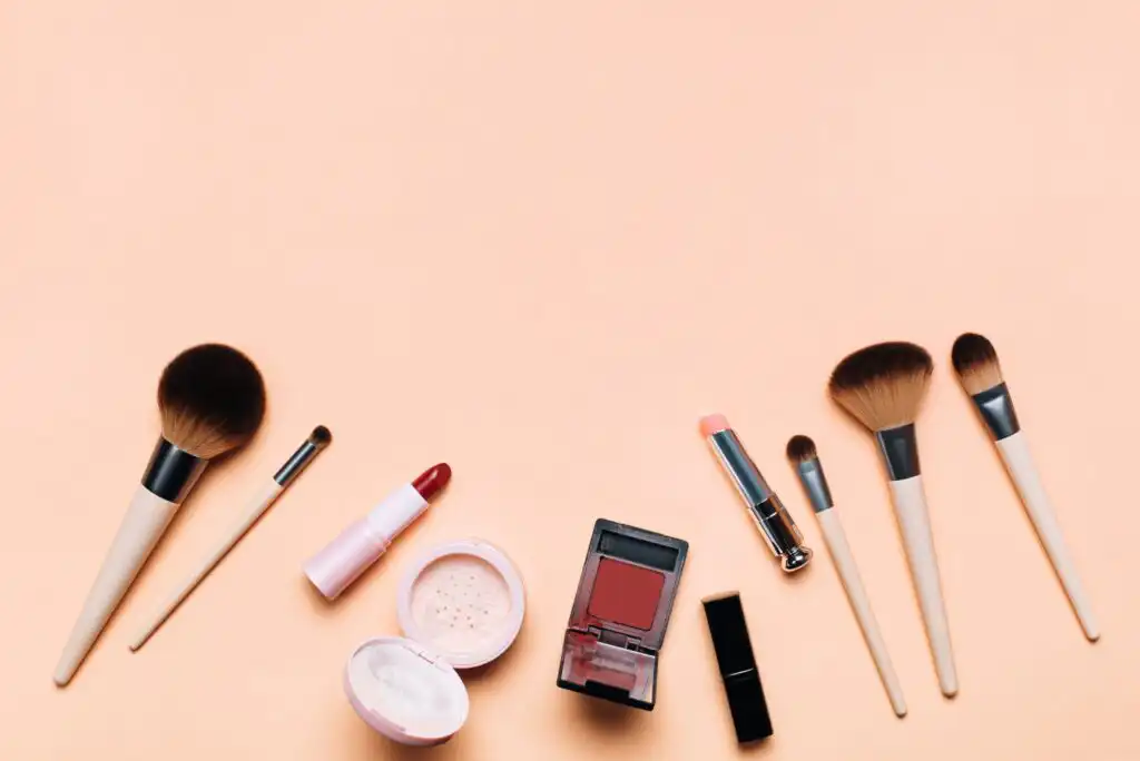 Various makeup products, including brushes, lipstick, blush, powder, and compact, are arranged neatly on a peach-colored background with empty space in the center.