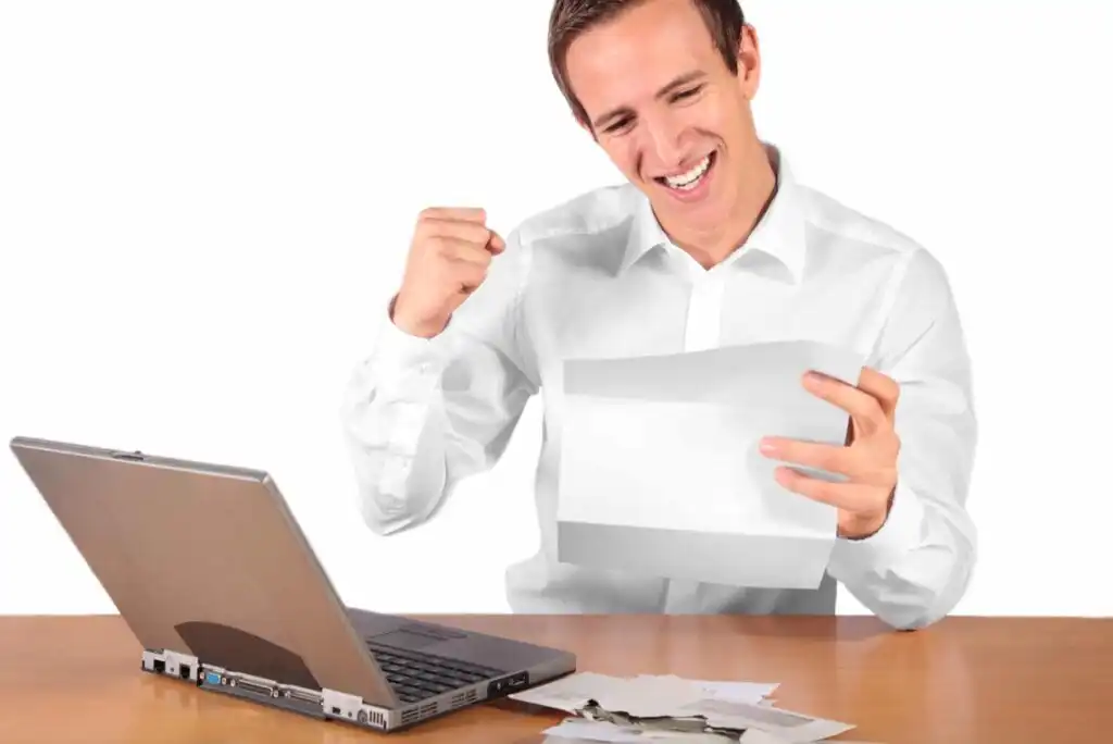 A man sitting at a desk with a laptop, smiling excitedly and raising his fist while holding a piece of paper, appearing pleased with the news or results he has received.