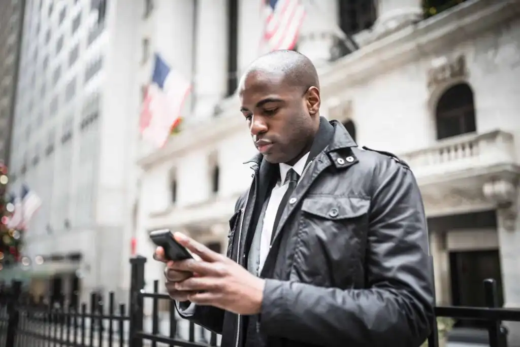 A man in a dark jacket and tie stands outside near a building with flags, looking at his smartphone. The background features columns and an iron fence, suggesting an urban or financial district.