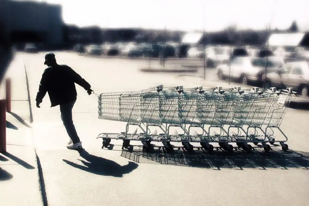 A person pushes a long line of empty shopping carts across a sunny parking lot, casting a shadow on the pavement. The background shows parked cars and a large building.