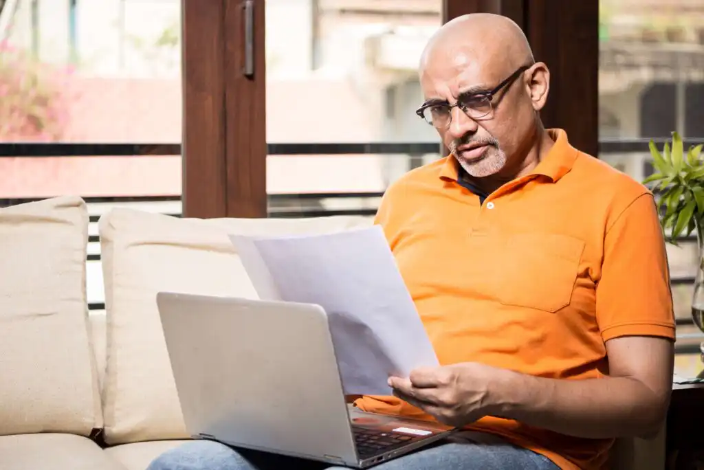 A man in an orange polo shirt sits on a couch with a laptop on his lap, reading a document with a focused expression. He is in front of a window with sunlight streaming in.