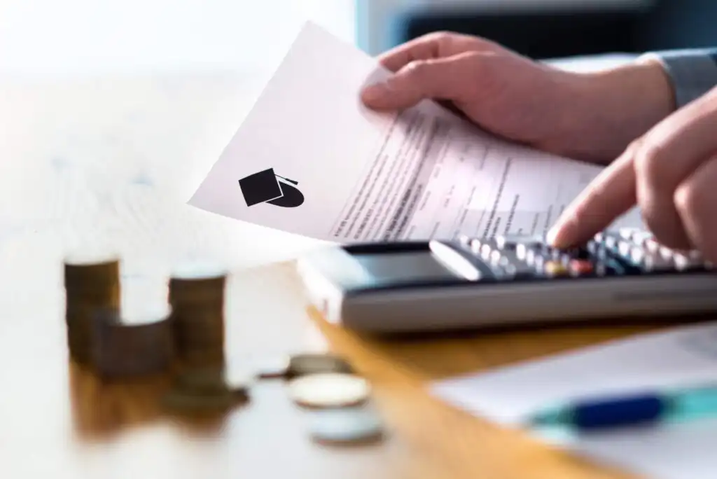 A person holds a paper with a graduation cap icon while using a calculator, with coins and bills stacked on a wooden table, representing financial planning for education.
