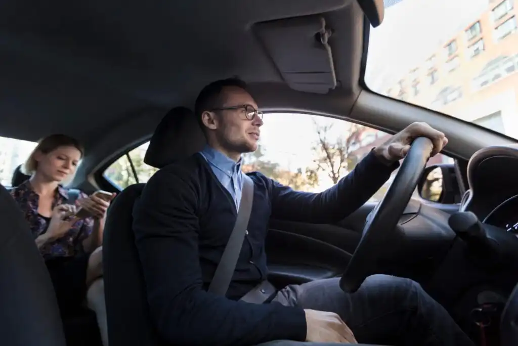 A man wearing glasses drives a car while a woman sits in the back seat looking at her phone. Both are wearing seatbelts, and city buildings are visible through the window.