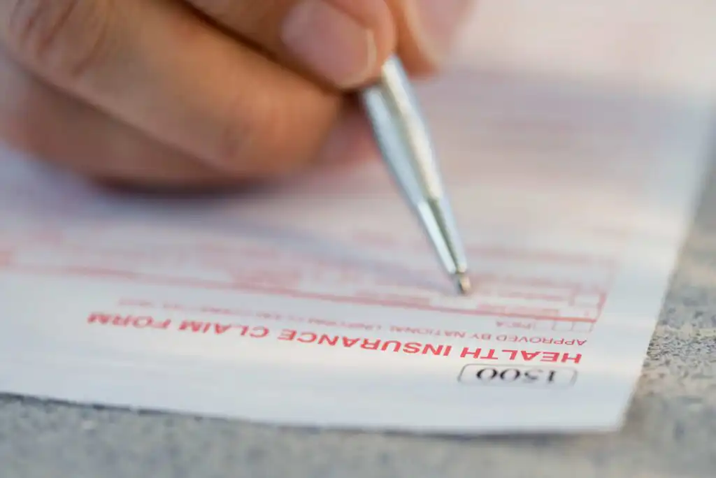 A close-up of a hand holding a pen and filling out a health insurance claim form. The words HEALTH INSURANCE CLAIM FORM are clearly visible in red text on the paper.