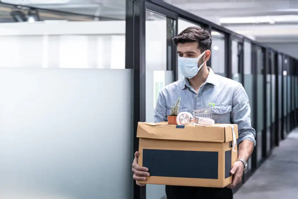 A man wearing a face mask carries a box of personal belongings, including a plant and a clock, while walking through an office corridor with glass partitions.