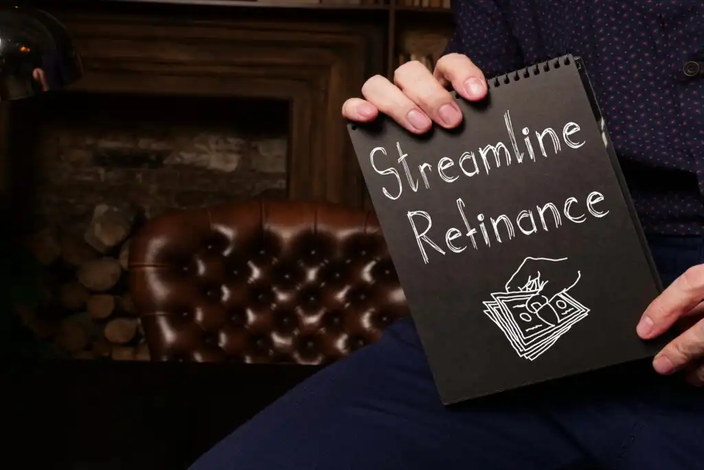A person holds a black notebook with Streamline Refinance and a drawing of money on the cover, sitting on a leather chair with a fireplace and stacked firewood in the background.