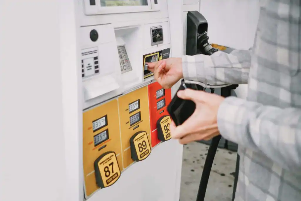 A person wearing a plaid shirt inserts a credit card into a gas pump, preparing to refuel their vehicle. The pump displays fuel options with octane ratings 87, 89, and 91.