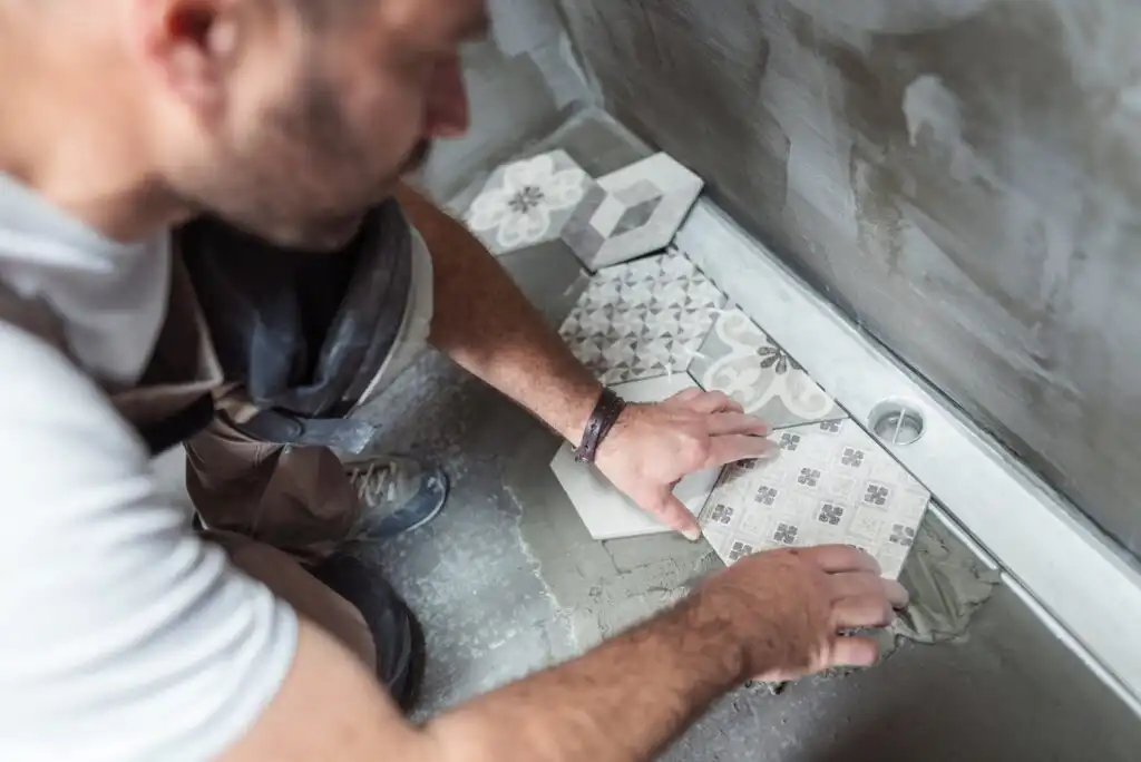 A person installing patterned ceramic tiles on a bathroom floor, carefully positioning one tile among others near a wall with fresh adhesive.