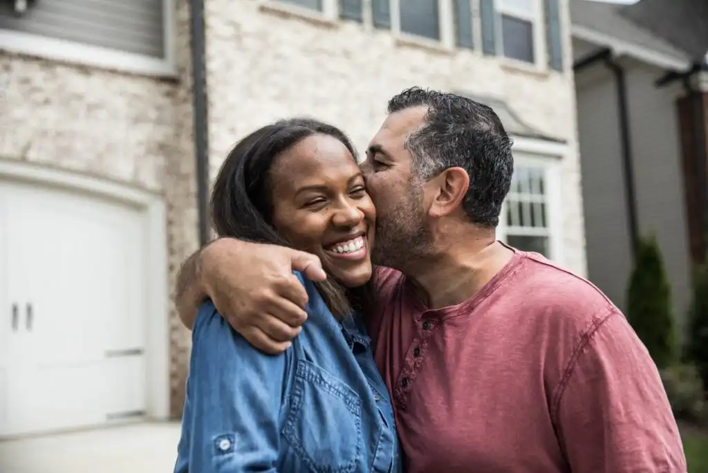 A man wearing a red shirt kisses a smiling woman in a blue shirt on the cheek, with his arm around her shoulders. They are standing outside in front of a brick house.