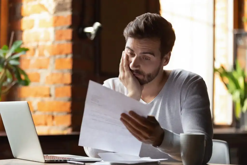 A man sits at a desk with a laptop and coffee cup, looking stressed as he holds and reads a piece of paper, resting his head on his hand in a modern, sunlit room.