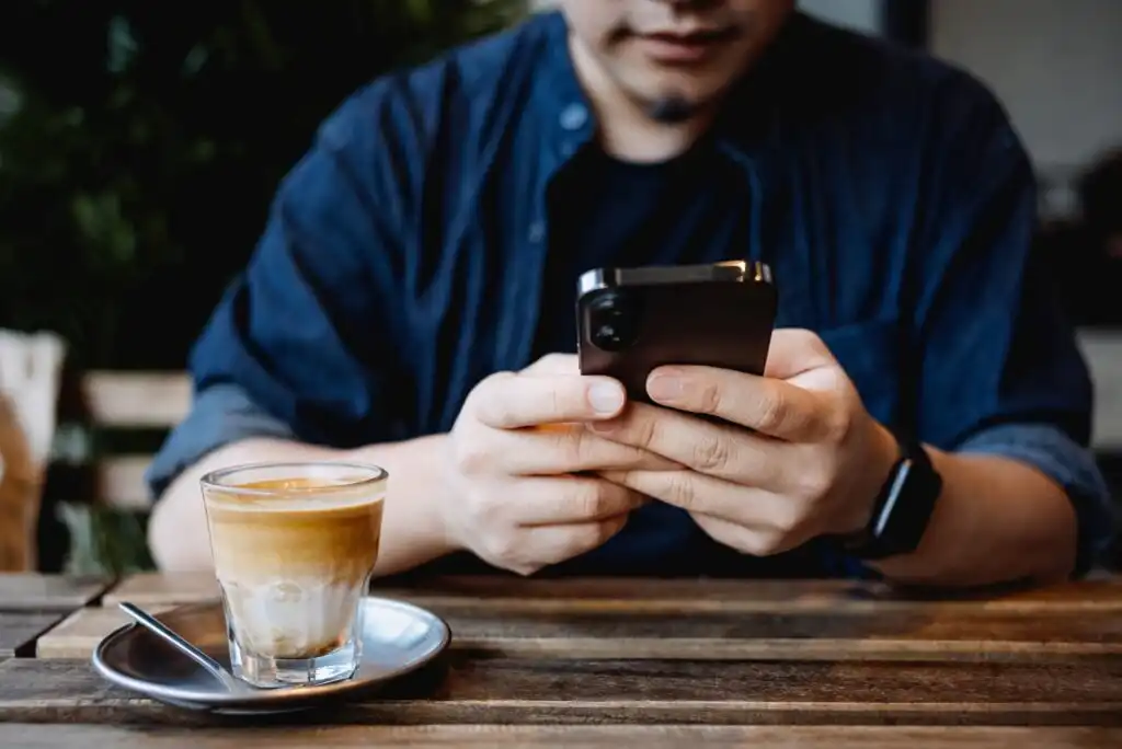 A person sitting at a wooden table using a smartphone, with a glass of coffee and a spoon on a saucer in front of them. The person is wearing a dark shirt and a wristwatch.