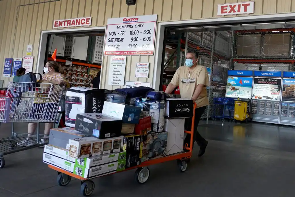 A man wearing a mask pushes a flatbed cart loaded with bulk goods out of a Costco store; store hours are posted above the entrance and exit doors. Other shoppers with carts are visible in the background.