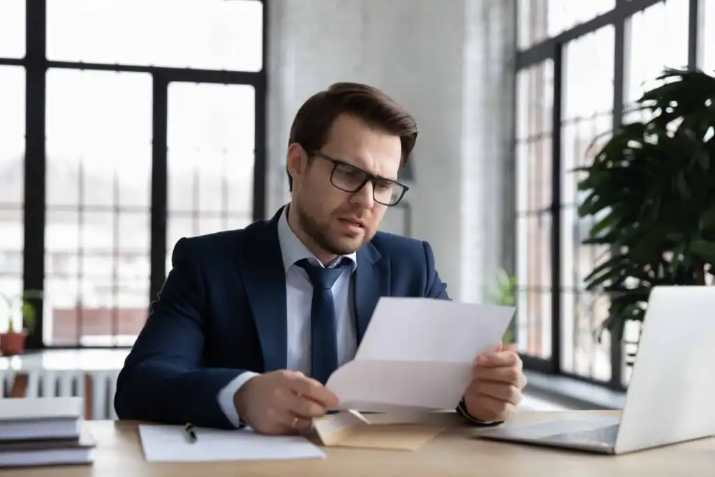 A man in a suit and glasses sits at a desk in an office, looking concerned while reading a document. A laptop and papers are on the desk, and large windows are in the background.
