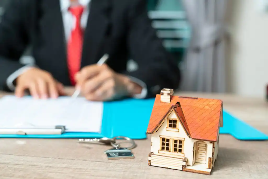 A small model house and a set of keys on a table with a person in a suit signing documents on a clipboard in the background, suggesting a real estate or home buying transaction.