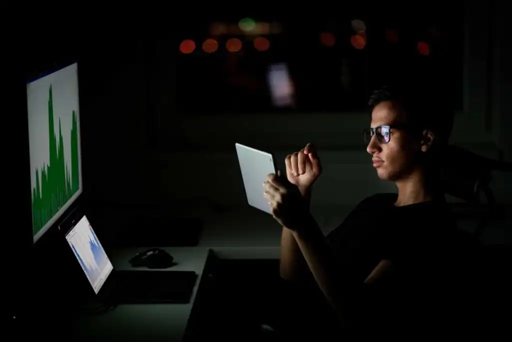A person wearing glasses is sitting in a dark room, looking at a tablet. Two computer screens on the desk display graphs with green and blue data. The only light comes from the devices.