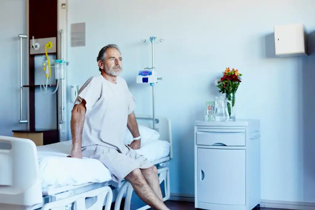 An older man in a hospital gown sits on the edge of a hospital bed in a bright, clean room. A bedside table with flowers, a glass of water, and medical equipment are visible nearby.