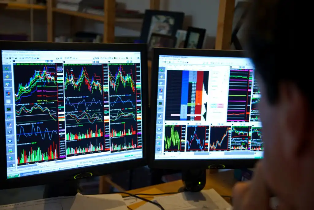 A person looks at two computer monitors displaying multiple colorful stock charts, graphs, and data analysis tools in a home or office setting.