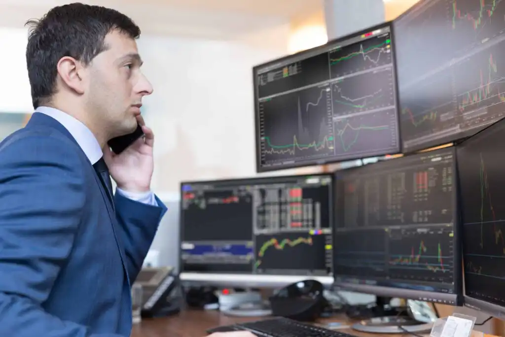 A man in a suit talks on the phone while looking at multiple computer monitors displaying financial charts and data in an office setting.