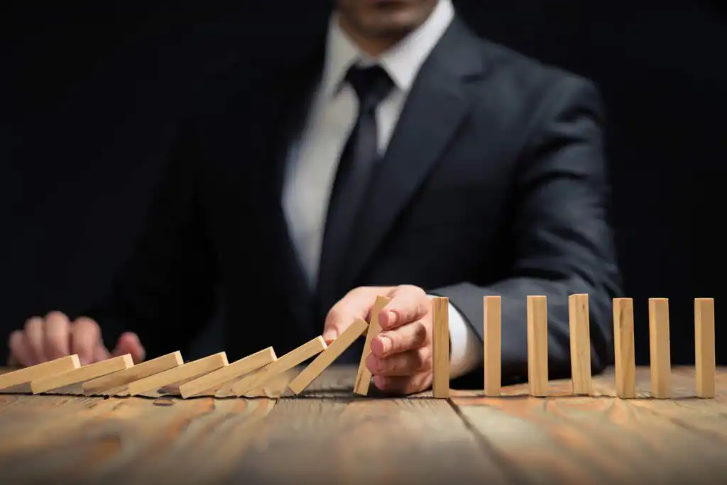A person in a suit stops a row of falling wooden dominoes with their hand on a wooden table, preventing the remaining upright pieces from toppling.