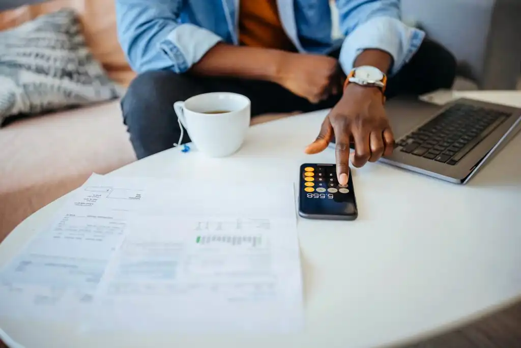 A person sits on a couch using a calculator on their phone at a white table covered with bills and documents, with a laptop and a cup of coffee nearby.