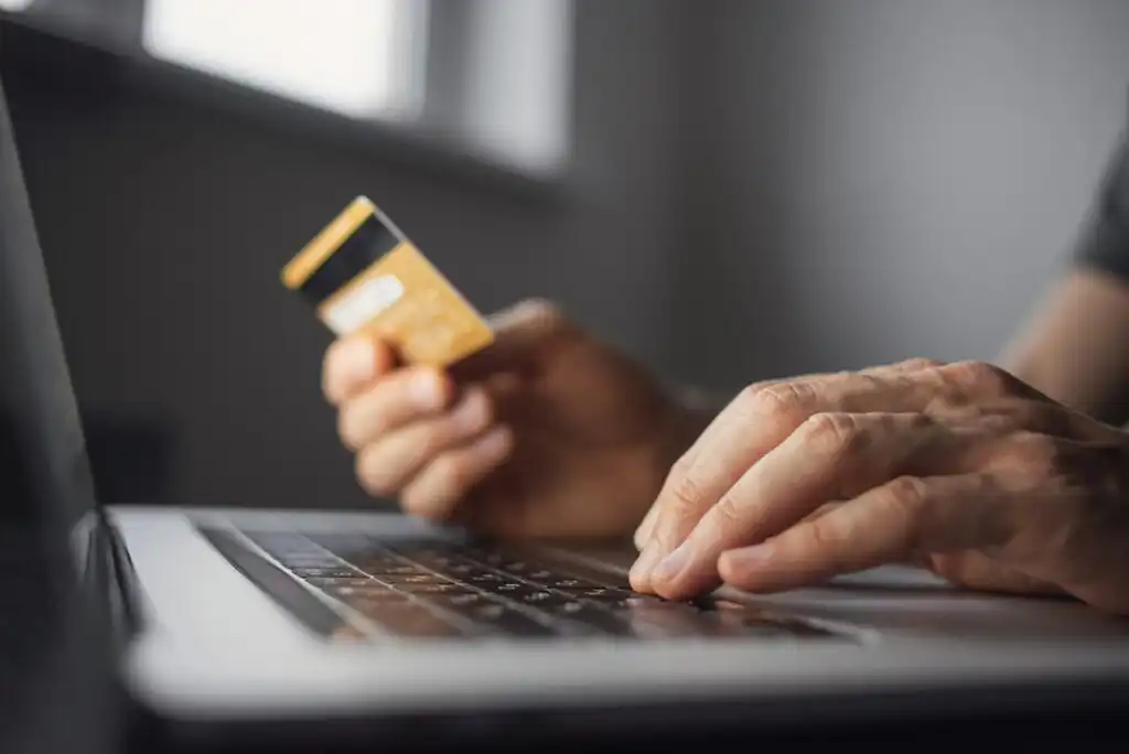 A person holds a credit card in one hand while typing on a laptop keyboard with the other, suggesting online shopping or digital payment.