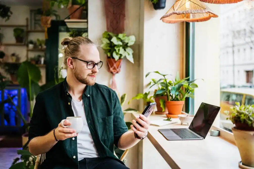A man with glasses and a beard sits by a window in a cozy café, holding a cup of coffee and looking at his phone. A laptop and several potted plants are on the wooden table in front of him.