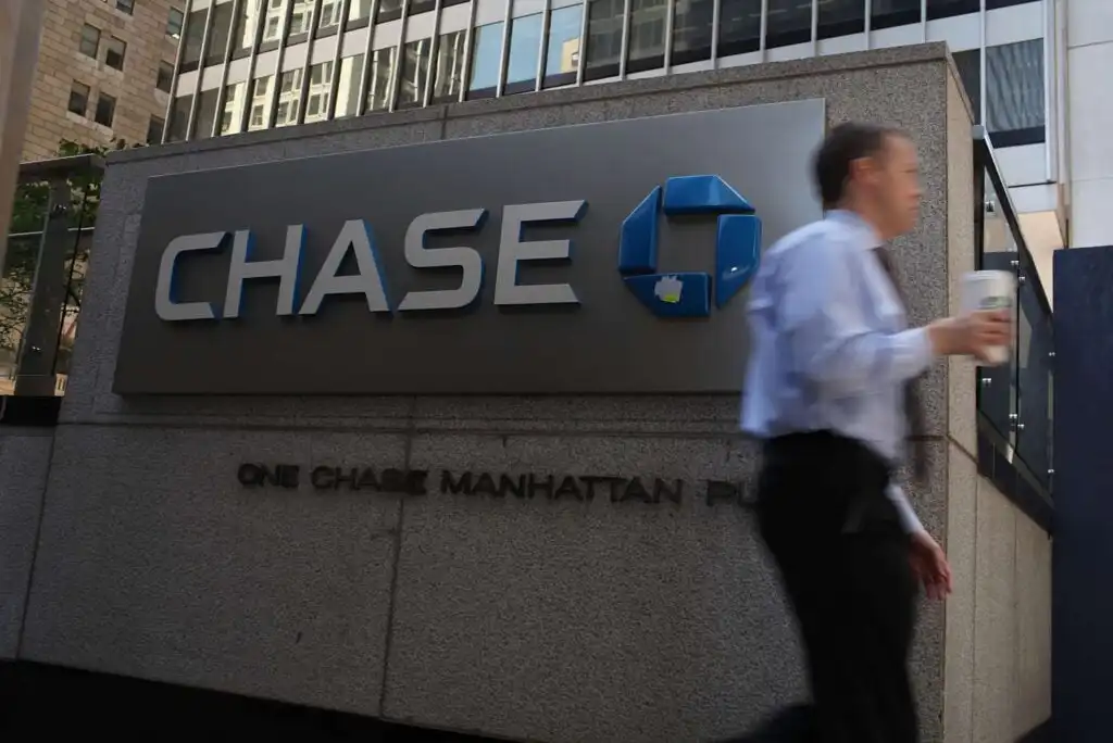 A person in business attire walks past a large Chase bank sign on a stone building, holding a coffee cup. Tall glass office buildings are visible in the background.