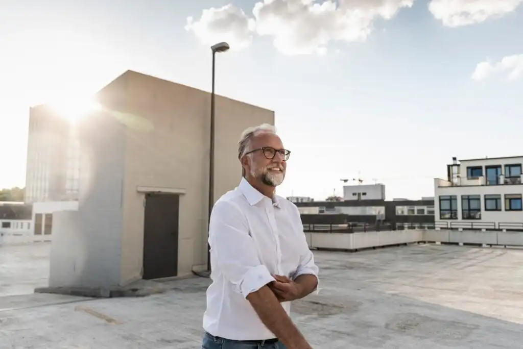 A smiling older man with glasses and a white shirt stands on a rooftop parking lot at sunset, rolling up his sleeve. Modern buildings and a clear sky with a few clouds are visible in the background.